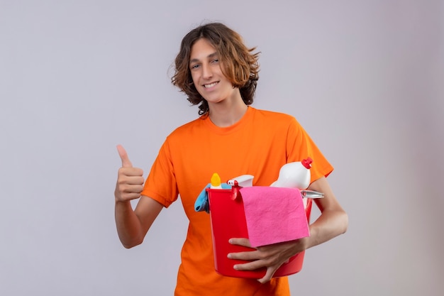 Jovem de camiseta laranja segurando um balde com ferramentas de limpeza, olhando para a câmera, sorrindo alegremente, mostrando os polegares para cima em pé sobre um fundo branco