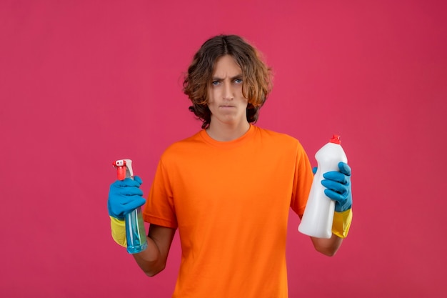 Jovem de camiseta laranja usando luvas de borracha segurando spray de limpeza e um frasco de material de limpeza olhando para a câmera descontente com o rosto carrancudo de pé sobre um fundo rosa