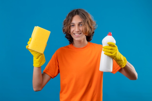 Jovem de camiseta laranja usando luvas de borracha segurando um frasco de material de limpeza e uma esponja olhando para a câmera sorrindo alegremente em pé sobre um fundo azul