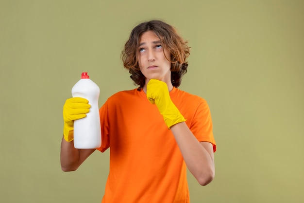 Jovem de camiseta laranja usando luvas de borracha segurando um frasco de material de limpeza em pé com a mão no queixo olhando para cima com uma expressão pensativa pensando sobre o fundo verde