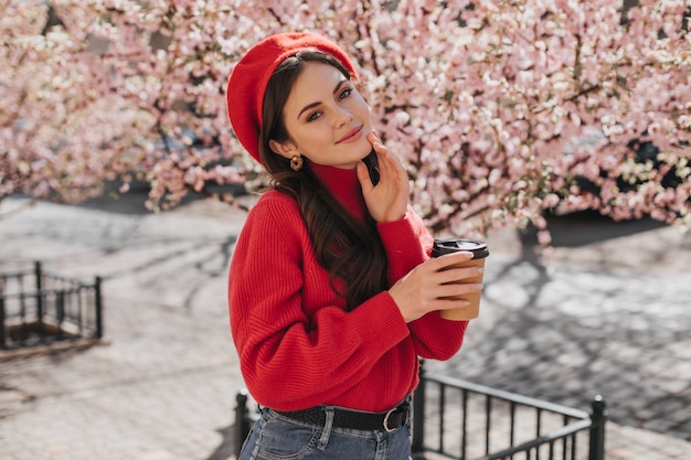 Jovem de camisola vermelha e chapéu está segurando uma xícara de café de papelão. jovem posando com um copo de chá e sorrindo sinceramente contra a flor de sakura