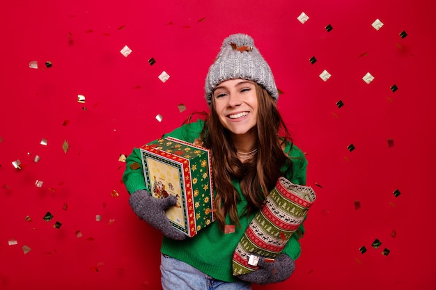 Jovem de energia atraente vestindo roupa de inverno segurando presentes de feriado sobre fundo vermelho isolado com confete, celebração, ano novo, aniversário, bom humor