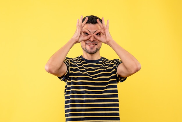 Jovem, de frente, com uma camiseta listrada em preto e branco fazendo binóculos com as mãos em um fundo amarelo isolado