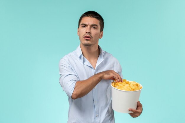 Jovem, de frente, segurando uma cesta com batata cips comendo e assistindo filme no cinema remoto solitário de parede azul claro