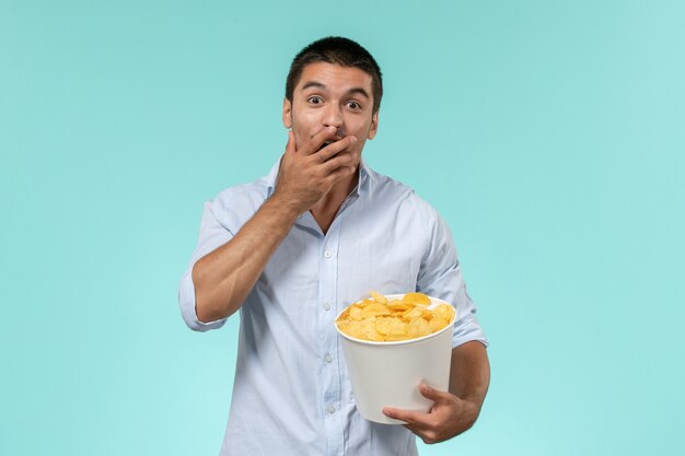 Foto grátis jovem, de frente, segurando uma cesta com batata cips comendo e assistindo filme no cinema remoto solitário de parede azul