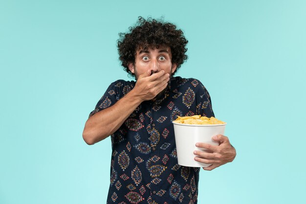 Jovem, de frente, segurando uma cesta com batatas cips e assistindo a um filme na parede azul do cinema.