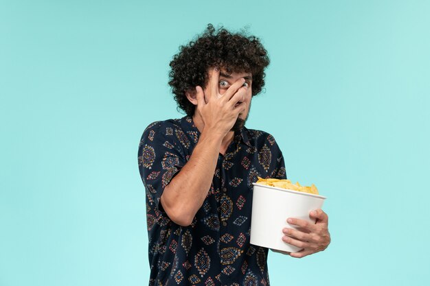 Jovem, de frente, segurando uma cesta com batatas cips e assistindo filme na mesa azul.