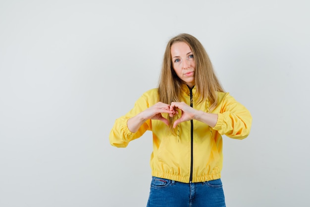 Jovem de jaqueta amarela e jeans azul, mostrando o gesto de coração e olhando encantadora, vista frontal.