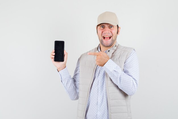 Jovem de jaqueta bege e boné segurando o telefone com uma mão e apontando para ele e olhando feliz, vista frontal.