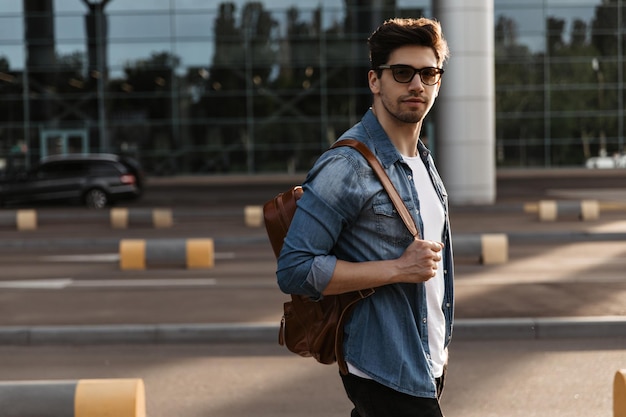 Jovem de jaqueta jeans de óculos de sol e camiseta branca olha para a câmera Linda morena segura mochila e posa do lado de fora