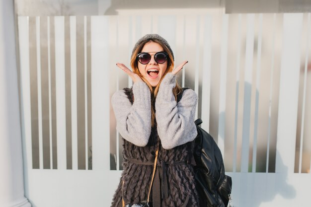 Jovem de óculos de sol da moda, aproveitando o bom tempo durante a caminhada matinal. Retrato ao ar livre de uma mulher animada em um suéter cinza carregando uma mochila preta e posando com uma expressão de surpresa.