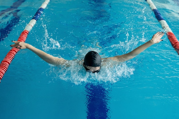 Jovem de óculos e boné natação estilo borboleta na piscina de corrida interior de água azul.