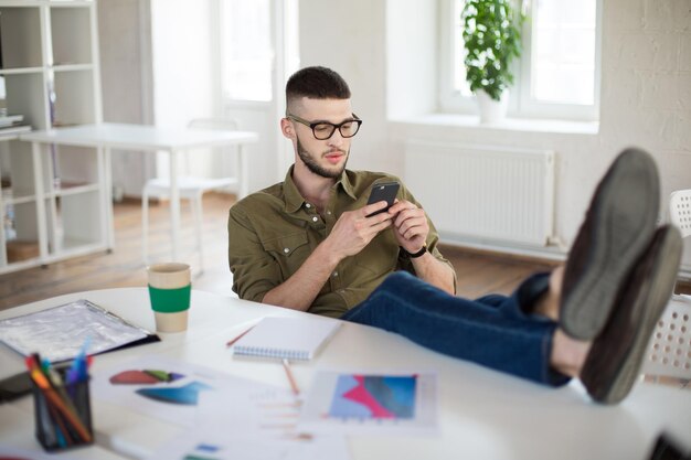 Jovem de óculos e camisa usando o celular enquanto está sentado na cadeira com as pernas na mesa no trabalho Empresário pensativo trabalhando no escritório moderno