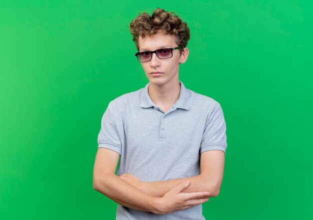 Jovem de óculos escuros vestindo uma camisa pólo cinza com rosto sério e mãos cruzadas em pé sobre uma parede verde
