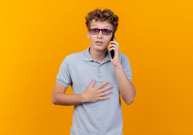 Jovem de óculos escuros, vestindo uma camisa pólo cinza, falando no celular, parecendo surpreso em pé sobre a parede laranja