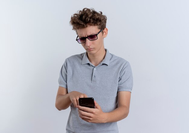 Jovem de óculos escuros, vestindo uma camisa pólo cinza, olhando para o celular do smartphone com uma cara séria em pé sobre uma parede branca
