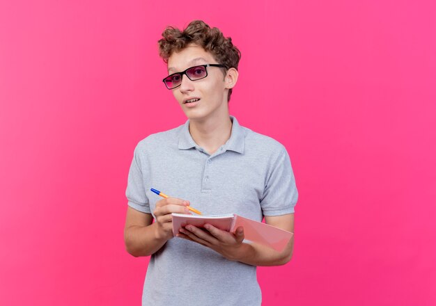 Jovem de óculos escuros vestindo uma camisa pólo cinza segurando um caderno com uma caneta feliz e positivo sorrindo rosa