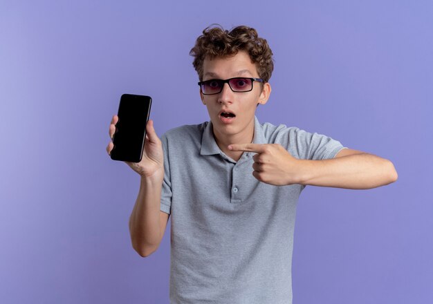 Jovem de óculos escuros vestindo uma camisa pólo cinza segurando um smartphone, apontando com o dedo indicador para ele, sendo surpreendido em pé sobre uma parede azul
