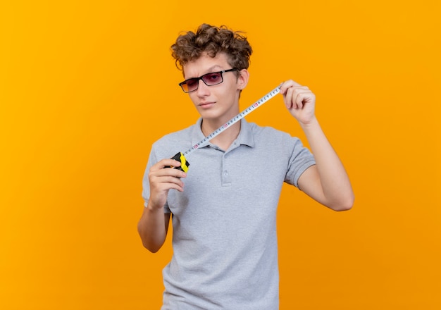 Jovem de óculos escuros vestindo uma camisa pólo cinza segurando uma fita métrica sorrindo feliz e positivo sobre laranja