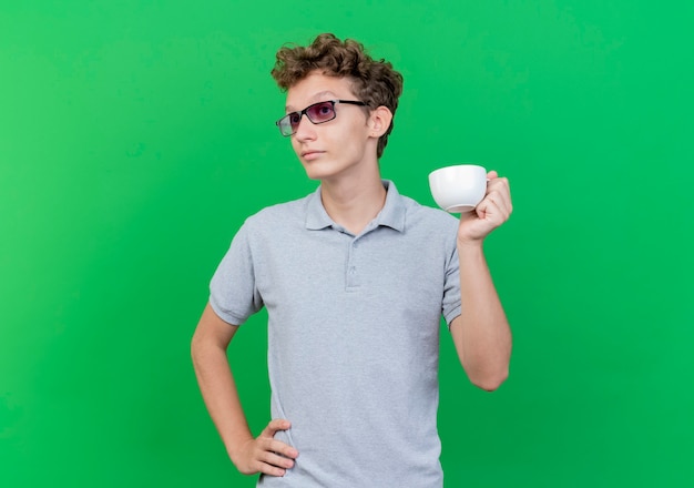 Jovem de óculos escuros, vestindo uma camisa pólo cinza segurando uma xícara de café, olhando para o lado, estando confuso e descontente em pé sobre uma parede verde