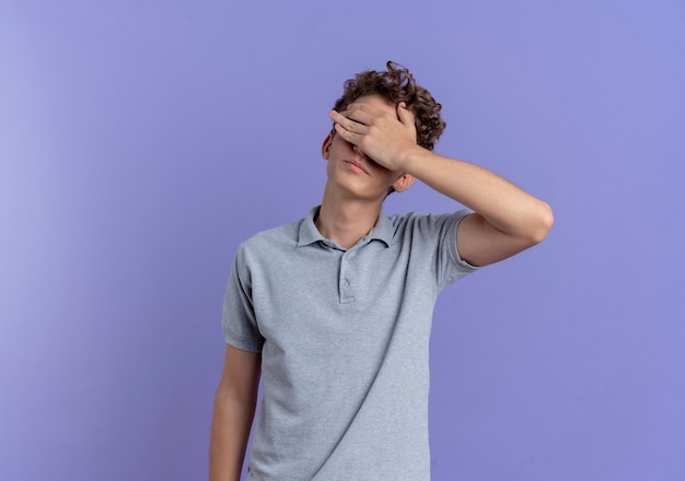 Jovem de óculos pretos usando uma camisa pólo cinza cobrindo os olhos com a mão cansada e entediada em pé sobre uma parede azul