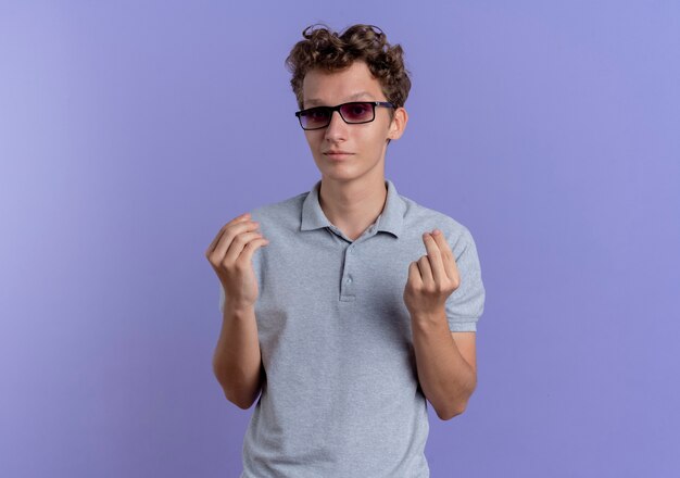 Jovem de óculos pretos usando uma camisa pólo cinza fazendo gesto de dinheiro esfregando os dedos em pé sobre a parede azul