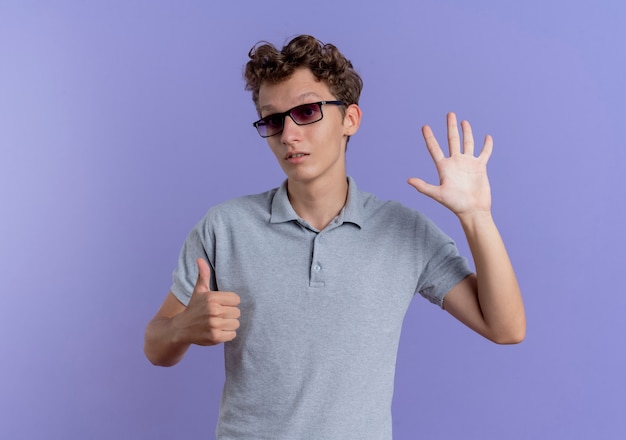 Jovem de óculos pretos vestindo uma camisa pólo cinza aparecendo e apontando para cima com os dedos número seis em pé sobre a parede azul