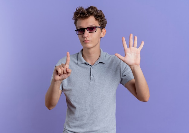 Jovem de óculos pretos vestindo uma camisa pólo cinza com rosto seriosu aparecendo e apontando para cima com os dedos número seis em pé sobre a parede azul