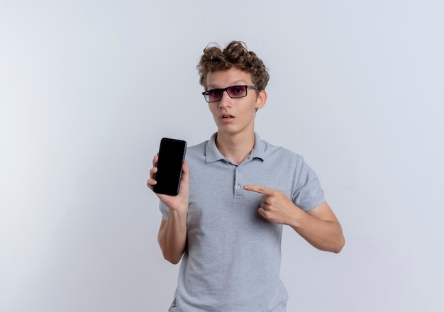 Jovem de óculos pretos, vestindo uma camisa pólo cinza, mostrando o smartphone apontando com o dedo para ele, sendo confuso em pé sobre uma parede branca