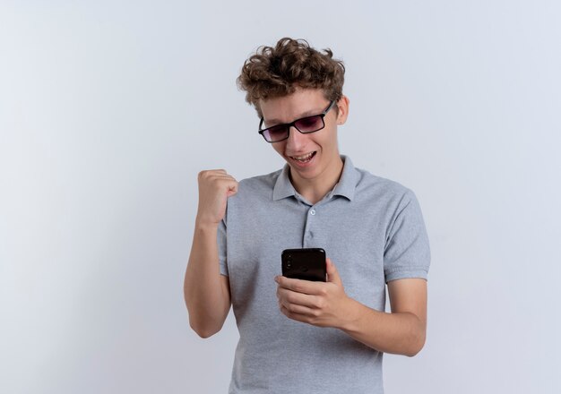 Jovem de óculos pretos, vestindo uma camisa pólo cinza, olhando para a tela do seu smartphone, o punho cerrado, feliz e animado em pé sobre uma parede branca