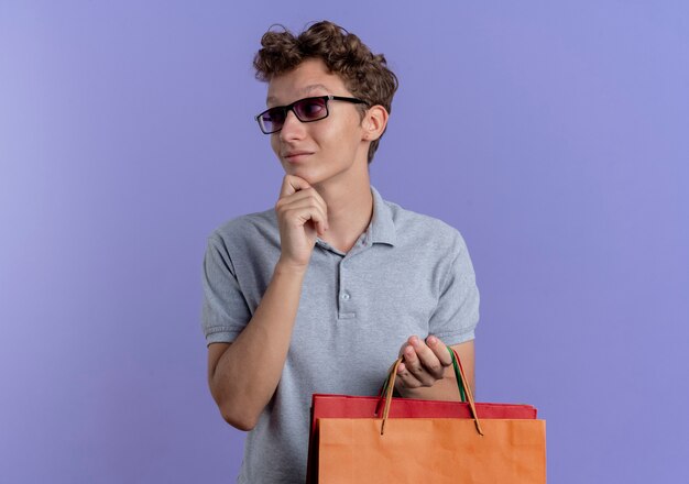 Jovem de óculos pretos vestindo uma camisa pólo cinza segurando sacolas de papel olhando para o lado com um olhar no queixo pensando em pé sobre uma parede azul