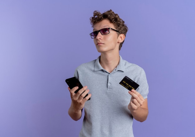 Jovem de óculos pretos, vestindo uma camisa pólo cinza segurando um smartphone e um cartão de crédito, olhando perplexo em pé sobre a parede azul