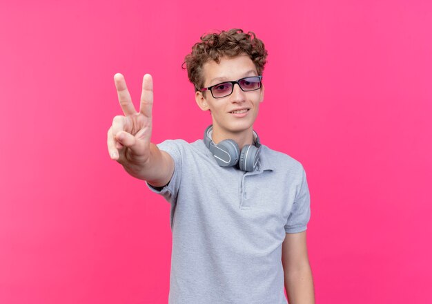 Jovem de óculos pretos, vestindo uma camisa pólo cinza, sorrindo e mostrando o sinal V feliz e alegre sobre rosa