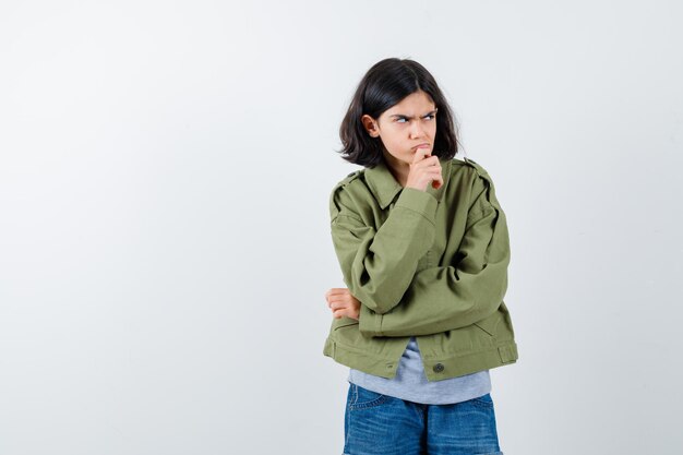 Jovem de pé em pose de pensamento de suéter cinza, jaqueta cáqui, calça jeans e olhando pensativa. vista frontal.