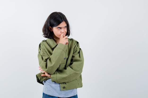 Jovem de pé em pose de pensamento de suéter cinza, jaqueta cáqui, calça jeans e olhando pensativa. vista frontal.