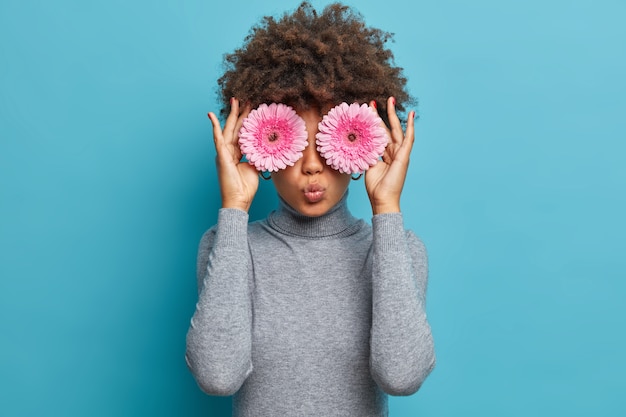 Jovem de pele escura cobre os olhos com lindas margaridas gérberas rosadas, brinca com as flores favoritas, desfruta de um aroma agradável, mantém os lábios fechados