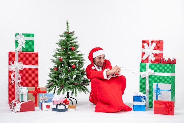 Jovem de rosto amargo vestido de Papai Noel com presentes e uma árvore de Natal decorada sentado no chão sobre um fundo branco