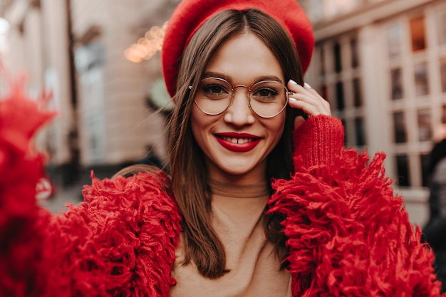 Jovem de roupa vermelha brilhante e chapéu faz selfie Senhora de óculos com batom vermelho posando contra o pano de fundo do belo edifício