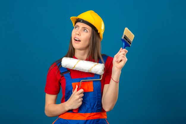 Jovem de uniforme de construção e capacete de segurança amarelo, pensando uma expressão pensativa de idéia segurando o rolo de pintura e escova de pé sobre fundo azul