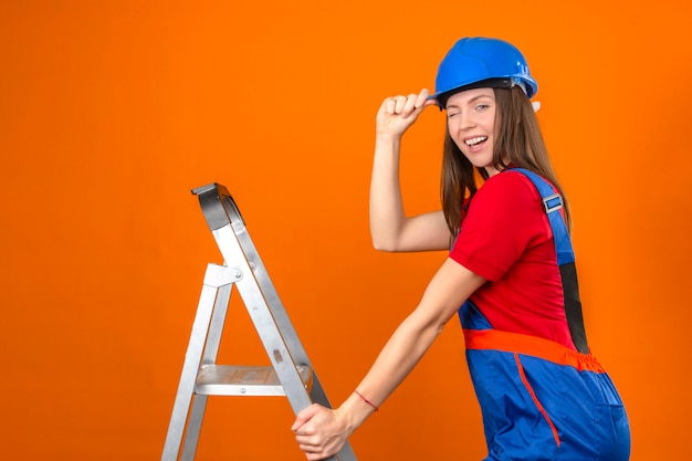 Jovem de uniforme de construção e capacete de segurança azul na escada piscando, olhando para a câmera no fundo laranja isolado