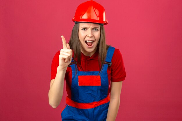 Jovem de uniforme de construção e capacete de segurança vermelho, apontando o dedo para cima com grande idéia saiu olhando para a câmera no fundo rosa escuro