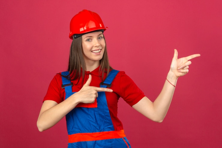 Foto grátis jovem de uniforme de construção e capacete de segurança vermelho sorrindo olhando para a câmera e apontando com a mão e o dedo para o lado de pé no fundo rosa escuro