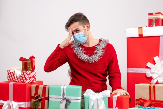 Jovem decepcionado com uma máscara sentado em frente aos presentes de natal