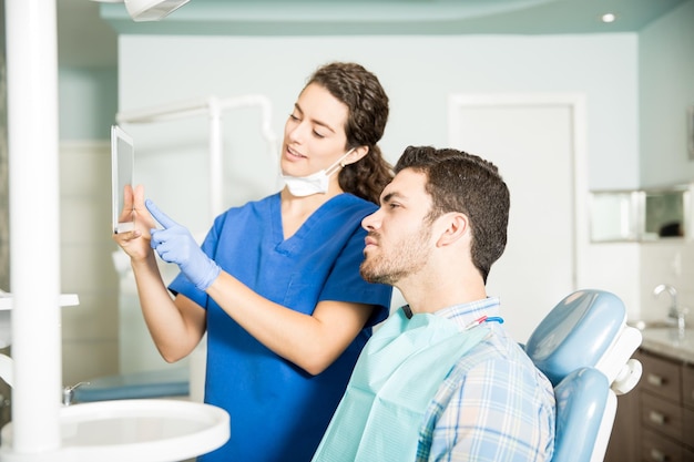 Jovem dentista mostrando tablet digital para paciente do sexo masculino durante o tratamento na clínica odontológica