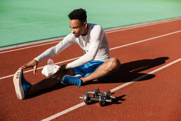 Jovem desportista afro-americano atraente, estendendo-se na pista de corrida no estádio da cidade