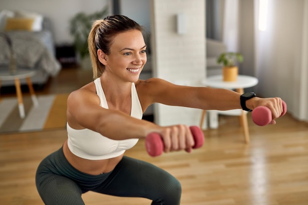 Jovem desportista feliz fazendo agachamentos enquanto se exercita com pesos de mãos na sala de estar