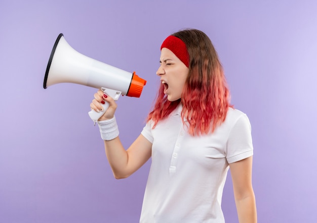 Jovem desportiva gritando para o megafone com expressão agressiva em pé sobre a parede roxa