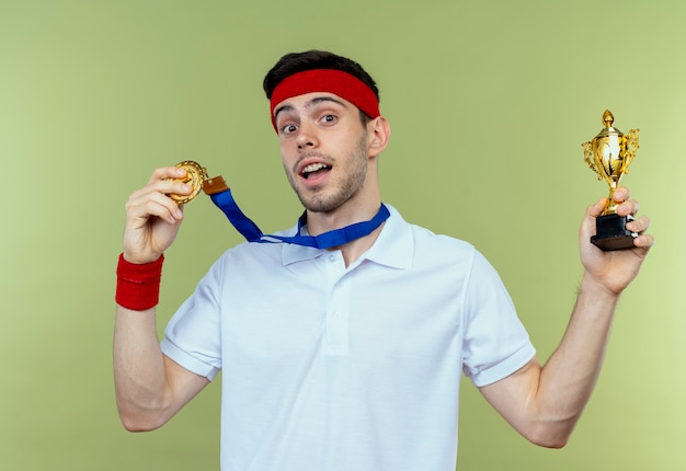 Jovem desportivo com bandana e medalha de ouro pendurada no pescoço segurando seu troféu, feliz e animado em pé sobre fundo verde