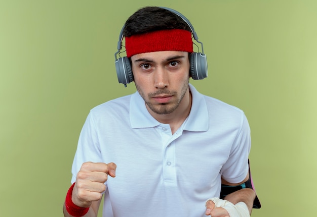 Foto grátis jovem desportivo com fita para a cabeça com fones de ouvido e pulseira de smartphone cerrando o punho com cara séria sobre verde