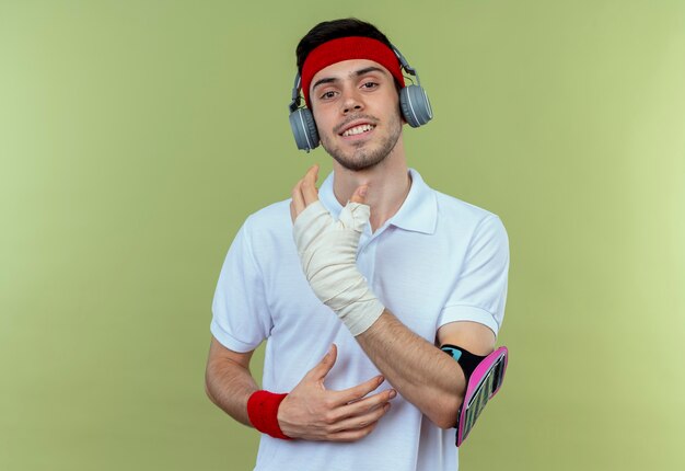 Jovem desportivo com fita para a cabeça com fones de ouvido e pulseira de smartphone sorrindo com o rosto feliz sobre o verde